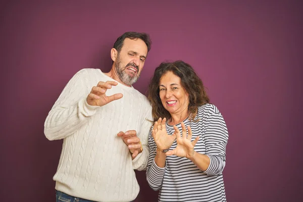 Beautiful middle age couple wearing winter sweater over isolated purple background disgusted expression, displeased and fearful doing disgust face because aversion reaction. With hands raised. Annoying concept.