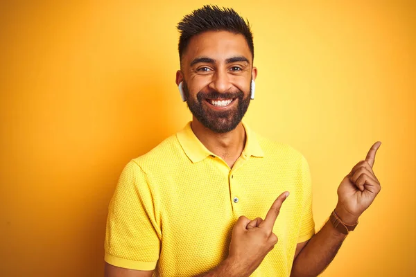 Young indian man listening to music using earphones standing over isolated yellow background smiling and looking at the camera pointing with two hands and fingers to the side.