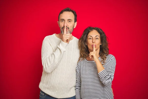 孤立した赤い背景の上に冬のセーターを着て美しい中年のカップルは 唇に指で静かにすることを求めています 沈黙と秘密の概念 — ストック写真