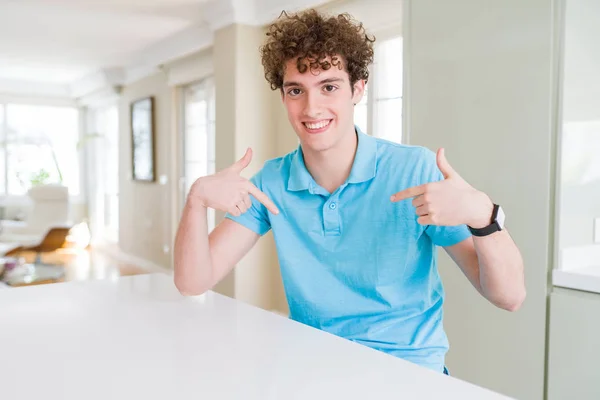 自宅でカジュアルな青いTシャツを着た若いハンサムな男は 笑顔に自信を持って見て 誇りに思って幸せな指で自分自身を指差す — ストック写真
