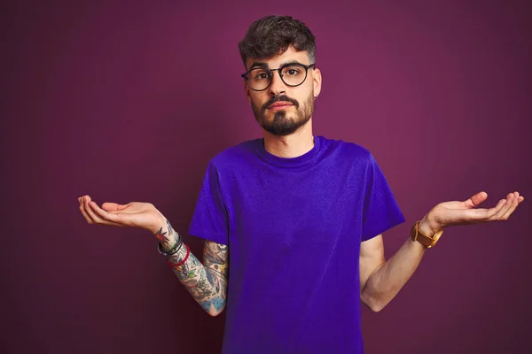 Tシャツとメガネをかけたタトゥーをした若者が 孤立した紫色の背景の上に立ち 腕と手を上げて混乱した表情を浮かべていた 疑いの概念 — ストック写真