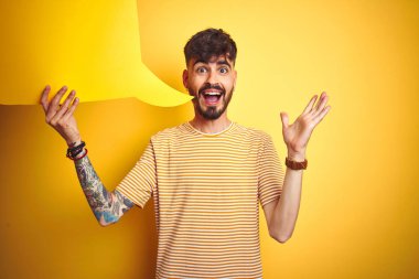 Dövme tutan konuşma balonu ile genç adam izole sarı arka plan üzerinde duran çok mutlu ve heyecanlı, kazanan ifade büyük bir gülümseme ile çığlık zafer kutluyor ve ellerini kaldırdı