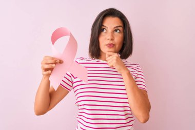 Genç güzel kadın izole pembe arka plan üzerinde duran kanser kurdele tutan ciddi yüz soru hakkında düşünme, çok karışık bir fikir