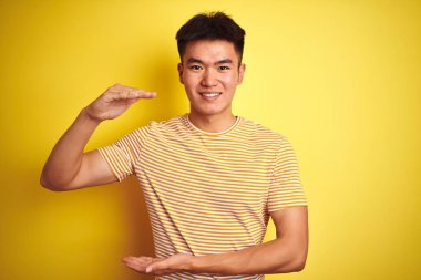 Genç Asyalı Çinli adam büyük ve büyük boy işareti, ölçmek sembolü gösteren eller ile izole sarı arka plan üzerinde ayakta t-shirt giymiş. Kameraya bakarak gülümsüyor. Ölçme konsepti.
