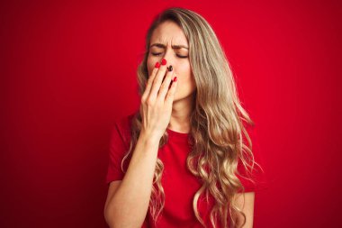 Kırmızı izole arka plan üzerinde ayakta temel t-shirt giyen genç güzel kadın el ile ağız kapsayan yorgun esneme. Huzursuz ve uykululuk.