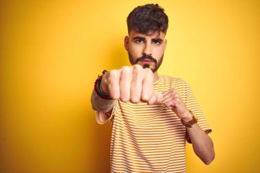 Dövme giyen genç adam izole sarı arka plan yumruk üzerinde duran çizgili t-shirt giyen mücadele, agresif ve öfkeli saldırı, tehdit ve şiddet