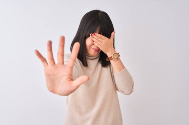 Genç güzel Çinli kadın el ile gözler kapsayan izole beyaz arka plan üzerinde balıkçı yaka kazak giyiyor ve üzücü ve korku ifadesi ile stop jest yapıyor. Utanç verici ve negatif kavram.