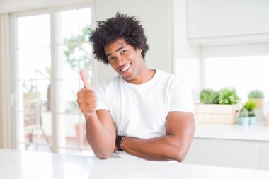 Genç afro-amerikan adam evde oturan rahat beyaz t-shirt giyen gururlu arıyorsunuz, yan jest kadar başparmak yapıyor gülümseyerek
