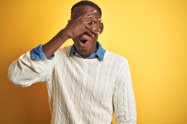 Kot gömlek ve beyaz kazak giyen Afrikalı Amerikan adam izole sarı arka planda şok kapsayan yüz ve gözleri el ile göz, utanmış ifade ile parmaklar bakarak.