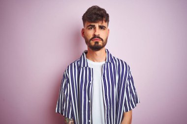 Dövmeli genç adam çizgili gömlek giyen izole pembe arka plan üzerinde ayakta depresif ve sıkıntı için endişe, kızgın ve korkmuş ağlıyor. Hüzünlü ifade.