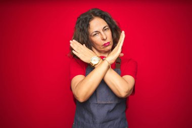 Orta yaş kıdemli kadın kırmızı izole arka planda apron üniforma giyiyor ret ifadesi geçiş kolları negatif işareti, öfkeli yüz yapıyor