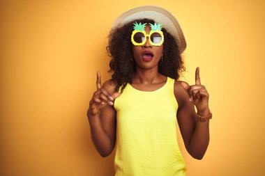 Izole sarı arka plan üzerinde komik ananas güneş gözlüğü giyen Afrikalı Amerikalı kadın şaşırttı ve yukarı bakarak ve parmakları ile işaret ve kolları kaldırdı şaşırttı.