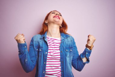 Güzel kızıl saçlı kadın kot gömlek giyiyor ve izole pembe arka plan üzerinde çizgili t-shirt şaşırttı ve silah kaldırdı ve gözleri ile başarı için hayret kutluyor kapalı. Kazanan kavramı.