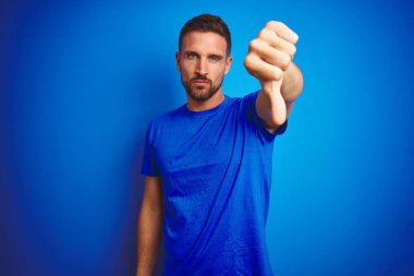 Genç yakışıklı adam mavi izole arka plan üzerinde rahat t-shirt giyen mutsuz ve kızgın gösteren reddi gösteren ve aşağı jest başparmak ile negatif. Kötü ifade.
