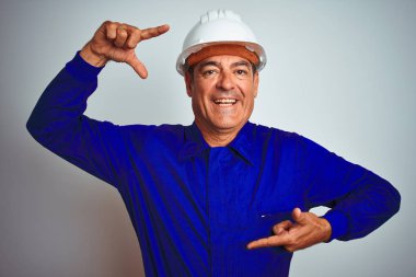 Yakışıklı orta yaş işçi adam üniforma ve kask üzerinde izole beyaz arka plan gülümseyen el ve parmaklarla mutlu yüz ile çerçeve yapıyor. Yaratıcılık ve Fotoğrafçılık konsepti.