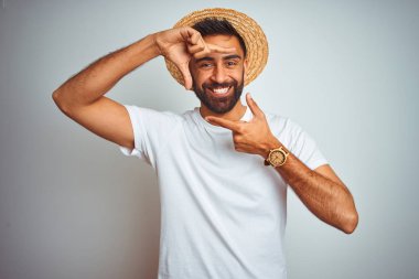 Genç Hint adam tatil giyen yaz şapka ayakta üzerinde izole beyaz arka plan gülümseyen yapmak çerçeve ile eller ve parmaklar ile mutlu yüz. Yaratıcılık ve Fotoğrafçılık konsepti.