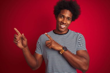 Afro saç ile genç Amerikan adam lacivert çizgili t-shirt üzerinde izole kırmızı arka plan gülümseyen ve kamera iki el ve parmak yan ile işaret bakıyor.