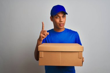Genç yakışıklı arap teslimat adam izole beyaz arka plan üzerinde duran bir kutu tutan mutlu yüz, bir numara ile bir fikir veya soru işaret parmak ile şaşırttı