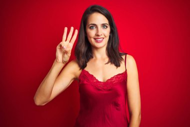 Kırmızı izole arka plan üzerinde seksi iç çamaşırı giyen genç güzel kadın gösteren ve parmak sayısı üç ile işaret kadar güven ve mutlu gülümseyen.