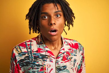 Tatil dreadlocks ile Afro adam izole sarı arka plan üzerine çiçek gömlek giyiyor sürpriz bir yüz ile şok korkuyor ve korku ifadesi ile heyecanlı korkmuş