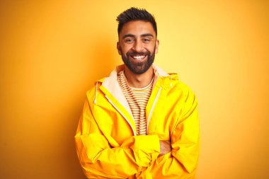 İzole sarı arka plan üzerinde ayakta yağmurluk giyen genç Hintli adam fotoğraf makinesi bakıyor çapraz silah ile gülümseyen yüz. Pozitif kişi.