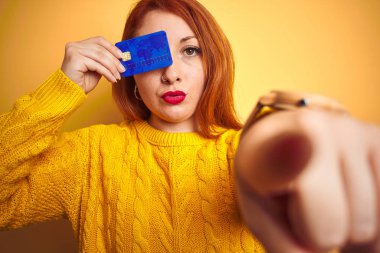 Kamera ve size, el işareti, ön emin jest parmak ile işaret sarı izole arka plan üzerinden kredi kartı tutan genç güzel Redhead kadın