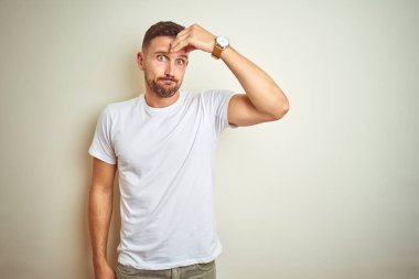İzole arka plan üzerinde rahat beyaz t-shirt giyen genç yakışıklı adam endişeli ve alnında el, sinir ve kriz için endişeli bir sorun hakkında vurguladı