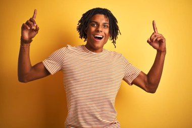 İzole sarı arka plan üzerinde ayakta çizgili t-shirt dikilen dreadlocks ile Afro adam hayran ve şaşırttı gülümsüyor ve parmaklar ve yükseltilmiş silah ile işaret.