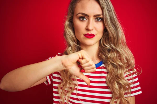 年轻美丽的女人穿着条纹T恤站在红色孤立的背景与愤怒的脸 负面的标志显示不喜欢与拇指向下 拒绝概念 — 图库照片