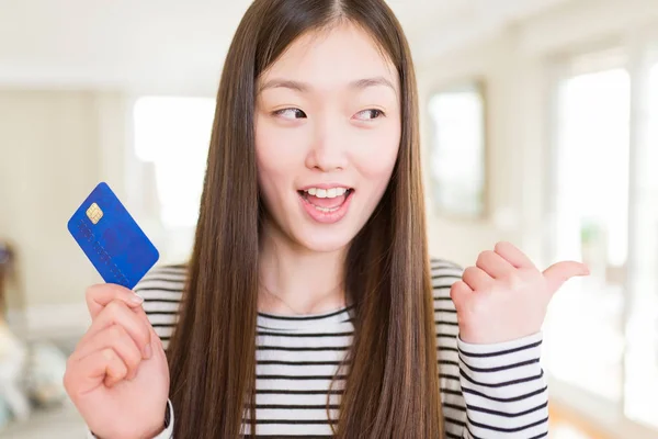 Beautiful Asian woman holding credit card pointing and showing with thumb up to the side with happy face smiling