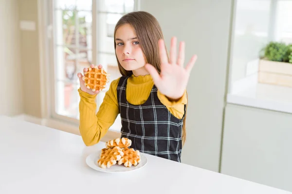 Beautiful young girl kid eating Belgian waffle pastry with open hand doing stop sign with serious and confident expression, defense gesture
