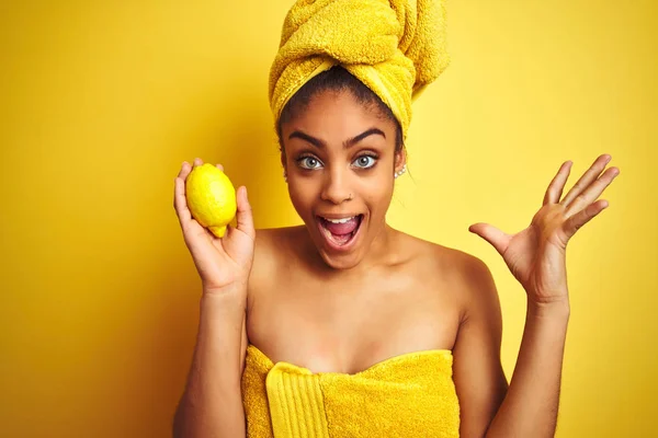 孤立した黄色の背景の上にレモンを持ってシャワーの後にタオルを身に着けているアフロアメリカの女性は非常に幸せで興奮し 大きな笑顔と手を上げて勝利を祝う勝者の表情 — ストック写真