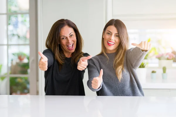 Beautiful family of mother and daughter together at home approving doing positive gesture with hand, thumbs up smiling and happy for success. Looking at the camera, winner gesture.
