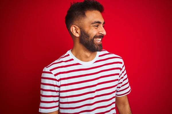 Young indian man wearing striped t-shirt standing over isolated red background looking away to side with smile on face, natural expression. Laughing confident.