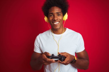 Afro oyun adam mutlu bir yüz ayakta ve dişleri gösteren kendinden emin bir gülümseme ile gülümseyerek izole kırmızı arka plan üzerinde joystick kulaklık kullanarak video oyunu oynuyor