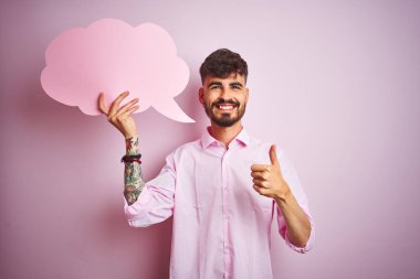 Dövme tutan bulut konuşma balonu büyük bir gülümseme ok işareti yapıyor mutlu izole pembe arka plan üzerinde duran genç adam, parmakları ile başparmak, mükemmel işareti