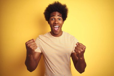 Afro saç, izole sarı arka plan üzerinde ayakta çizgili t-shirt giymiş Amerikan adam çok mutlu ve heyecanlı silah kaldırdı, gülümseyen ve başarı için çığlık ile kazanan jest yapıyor. Kutlama konsepti.