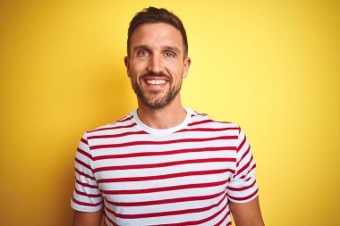 Yüzünde mutlu ve serin bir gülümseme ile sarı izole arka plan üzerinde Casual kırmızı çizgili t-shirt giyen genç yakışıklı adam. Şanslı kişi.
