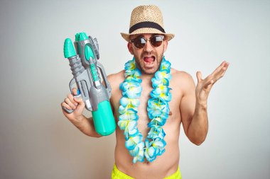 Genç adam yaz şapka giyen ve Hawaii Lei çiçek izole arka plan üzerinde su tabancası tutan çok mutlu ve heyecanlı, kazanan ifade zafer büyük gülümseme ile çığlık kutluyor ve el kaldırdı