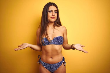 Genç güzel kadın tatilde bikini giyen izole sarı arka plan clueless üzerinde duran ve açık kollar ile karıştı, hiçbir fikrim yok kavramı.