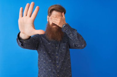 Genç kızıl saçlı İrlandalı adam elleri ile gözleri kaplayan izole mavi arka plan üzerinde duran çiçek yazlık gömlek giyen ve üzgün ve korku ifadesi ile stop jest yapıyor. Utanmış ve olumsuz kavram.