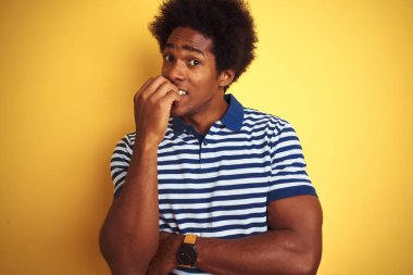 Afro saçlı Amerikan adam lacivert çizgili polo giyen izole sarı arka plan üzerinde duran stresli ve ağız tırnak ısırma elleri ile sinir görünümlü. Anksiyete sorunu.