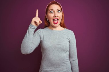Gri t-shirt giyen güzel kızıl saçlı kadın ve izole mor arka plan üzerinde diadem başarılı bir fikir ile parmak yukarı işaret. Çıkıldı ve mutlu. Bir numara.