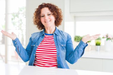 Evde kot ceket giyen kıvırcık saçlı orta yaş kıdemli kadın her iki el açık avuç gösteren gülümseyen, sunma ve reklam karşılaştırma ve denge