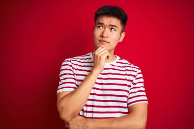 Genç Asyalı Çinli adam çizgili t-shirt üzerinde ayakta izole kırmızı arka plan üzerinde el çenesi ile soru hakkında düşünme, dalgın ifade. Düşünceli bir yüz ile gülümsüyor. Şüphe kavramı.