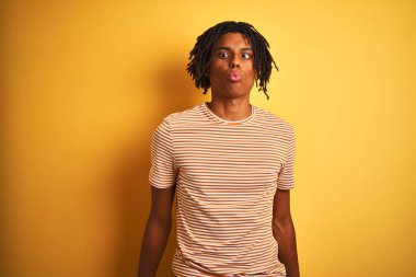 Dudak, çılgın ve komik jest ile balık yüz yapma izole sarı arka plan üzerinde ayakta çizgili t-shirt giyen dreadlocks ile Afro adam. Komik ifade.