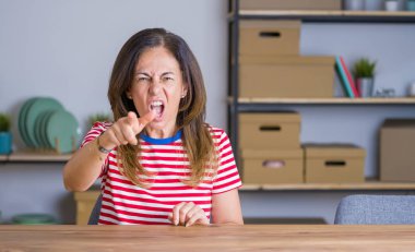 Orta yaş kıdemli kadın evde masada oturan memnun ve kamera, öfkeli ve öfkeli sizi sinirli işaret