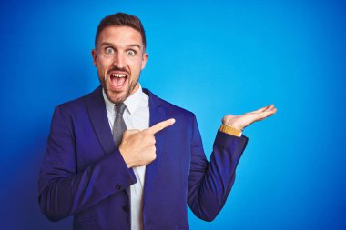 Mavi izole arka plan üzerinde genç yakışıklı iş adamı şaşırmış ve el ile sunmak ve parmak ile işaret ederken kameraya gülümsüyor.