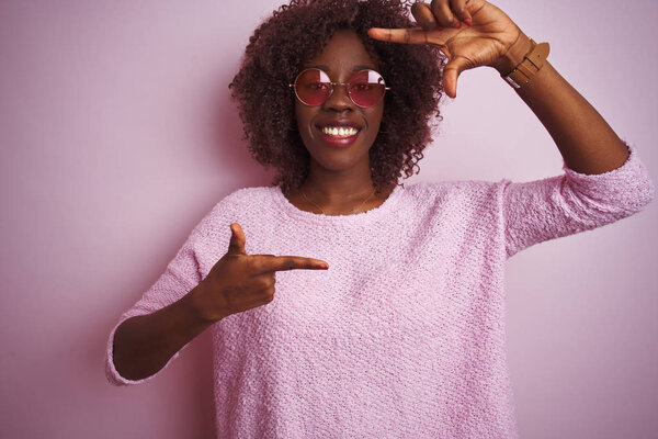 Young african afro woman wearing sweater and sunglasses over isolated pink background smiling making frame with hands and fingers with happy face. Creativity and photography concept.