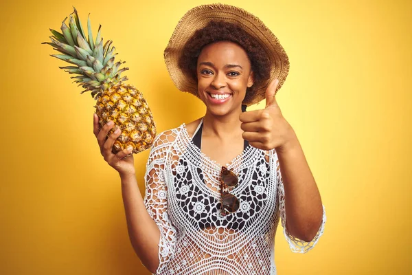 Young african american woman with afro hair holding pineaple over yellow isolated background happy with big smile doing ok sign, thumb up with fingers, excellent sign